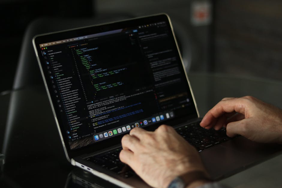 A close-up view of a person's hands typing on a laptop keyboard, displaying lines of code on the screen with green, blue, and white text on a dark background. The device is positioned on a black table